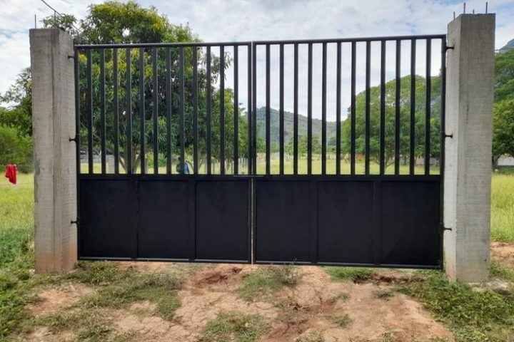 Portón negro de barrotes con paneles sólidos, montado entre postes de concreto en un predio rural